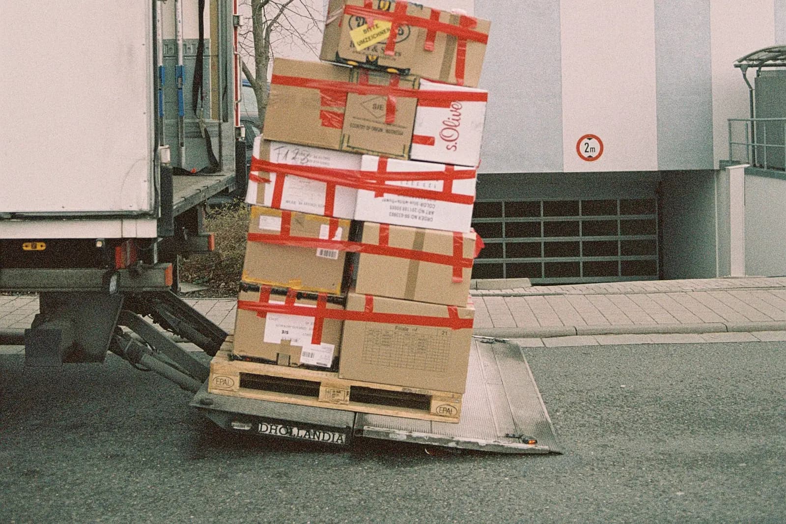 Palette de cartons sanglés, penchée sur le hayon d’un camion de livraison au bord d’un trottoir.