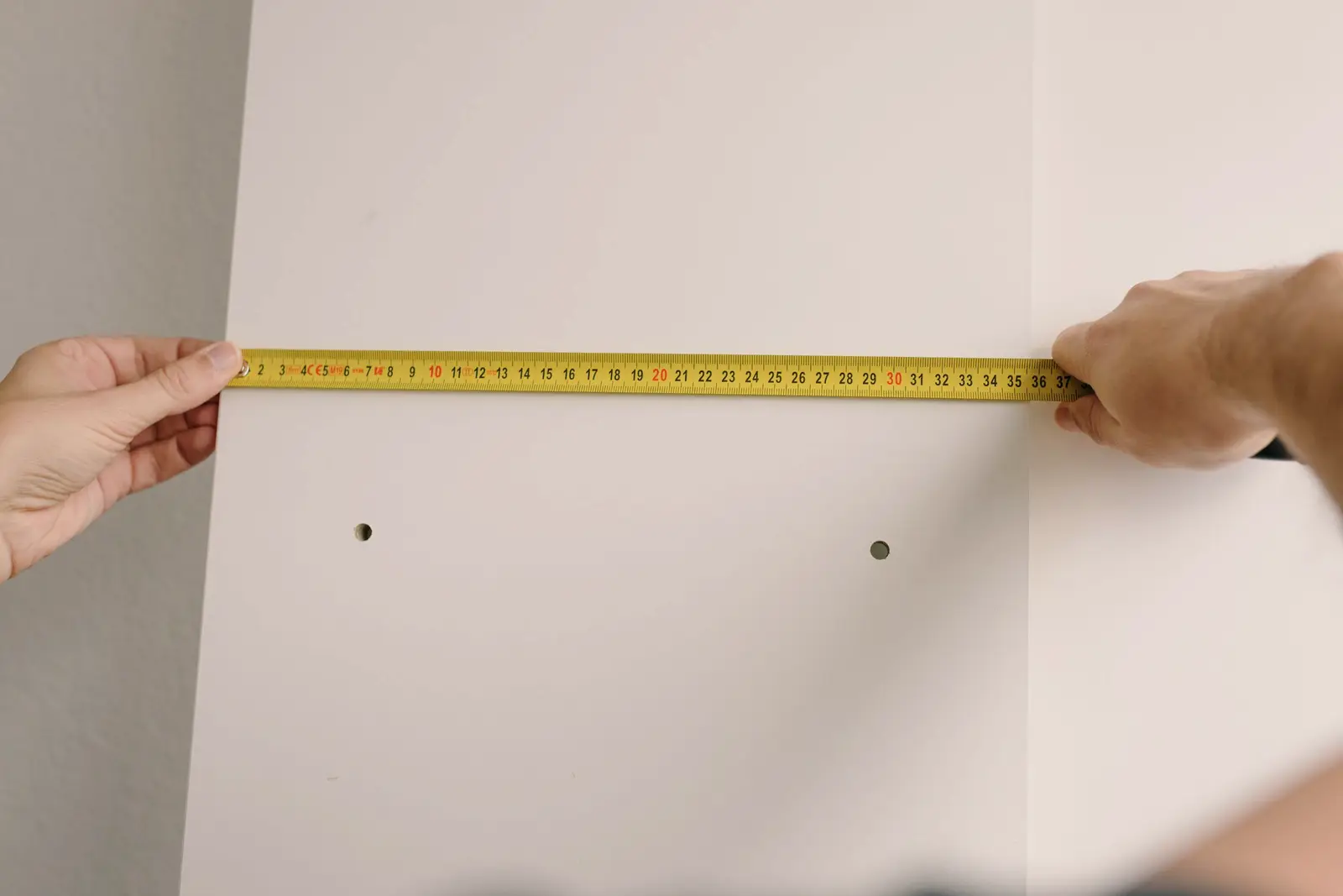 Deux mains tendant un mètre ruban en travers d’un panneau clair, au-dessus de deux points de fixation déjà percés.