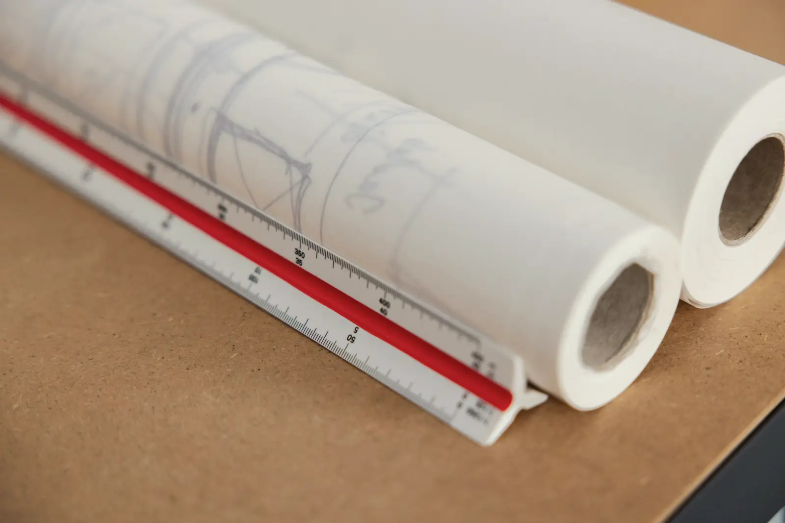 Deux rouleaux de plans posés à plat sur un plan de travail, un réglet d’architecte glissé contre eux.