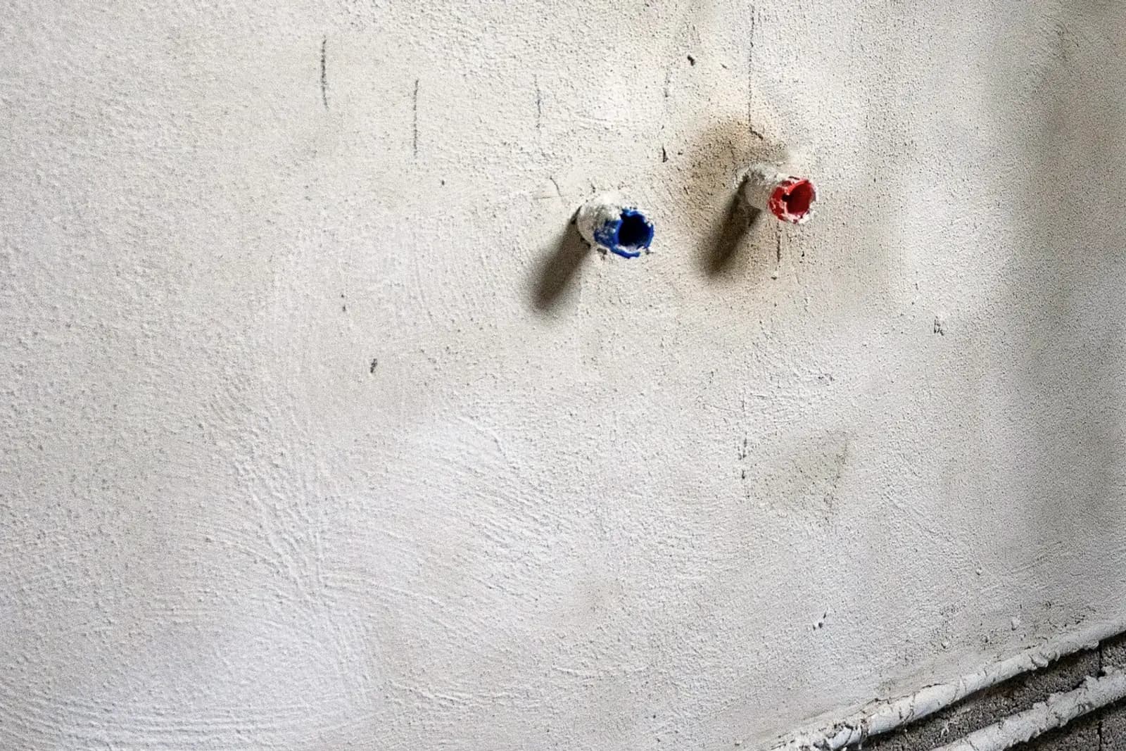 Deux attentes d’alimentation bouchées, rouge pour l’eau chaude et bleu pour l’eau froide, sur un mur enduit dont la saignée laisse encore voir les tubes au ras du sol.