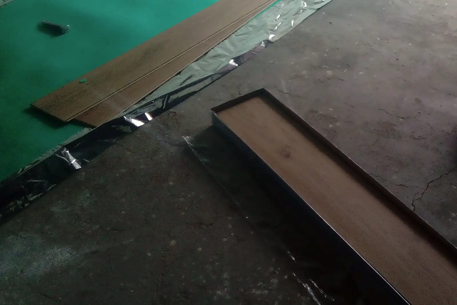 Sous-couche de parquet flottant déroulée sur une dalle brute fissurée, bordée d’un film aluminium, deux lames posées dessus et une troisième encore dans son carton.