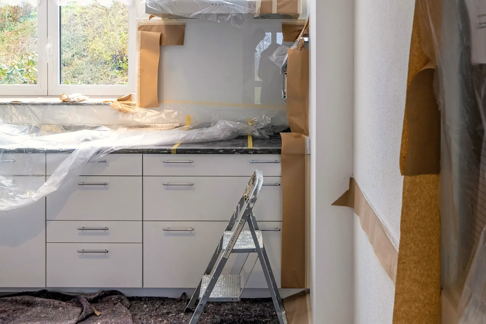 Rangée de meubles bas de cuisine blancs sous bâche plastique et papier kraft, escabeau et couverture de protection au sol, pendant des travaux.