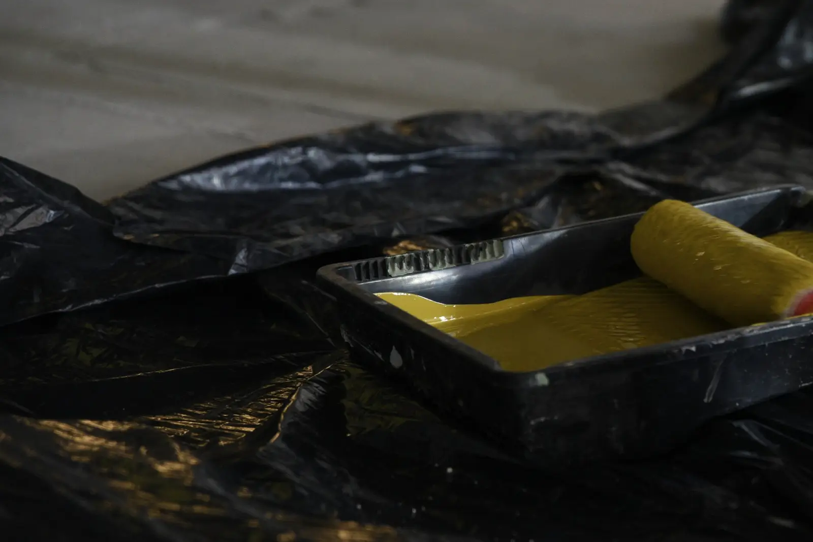 Bâche en polyéthylène étalée pour protéger le sol d’une dalle de béton, sous un bac à peinture jaune et son rouleau.