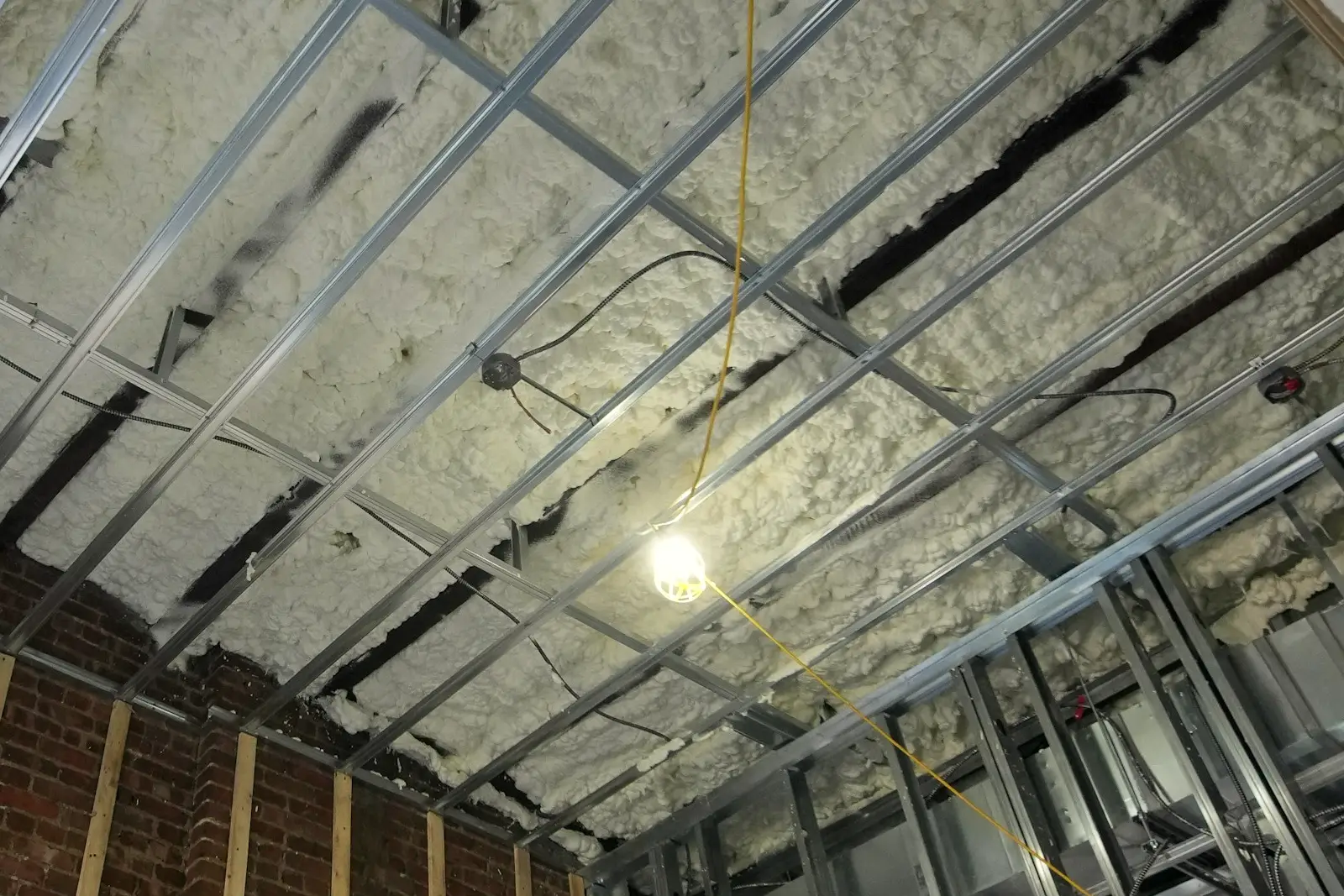 Ossature métallique d’un plafond encore ouvert sous un isolant projeté, câbles et boîte de point lumineux en attente, baladeuse de chantier allumée.
